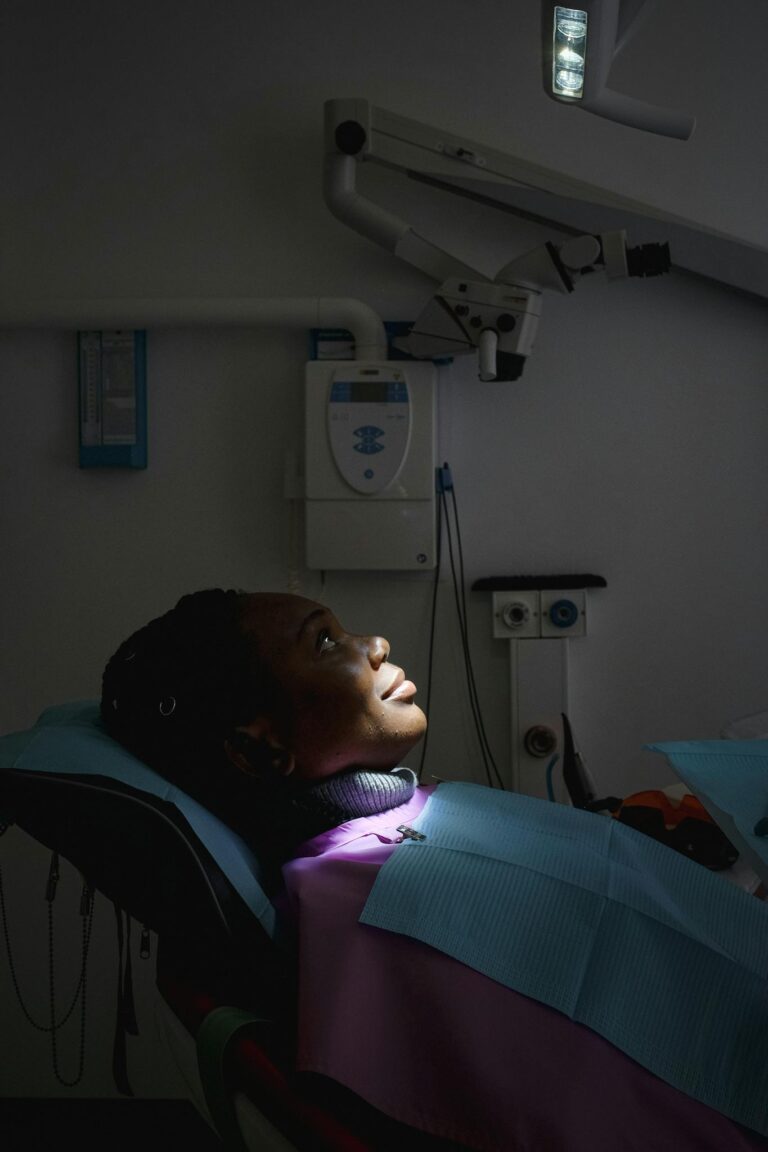Side view of young black woman in protective coat sitting in dental chair during X ray procedure in modern dark dental room and looking up